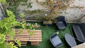 Une terrasse dotée de bancs en bois et de chaises noires, entourée de verdure et d’un mur en pierre.