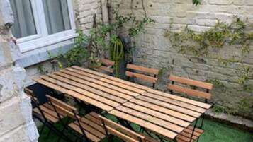 An outdoor patio with a wooden table and benches, surrounded by a brick wall and greenery.