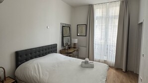 A hotel room with a large bed, a bedside table, a desk with a chair, a window with curtains, and a wall-mounted mirror. - Elegant 2-Bedroom Apartment in the Heart of Paris (Paris)
