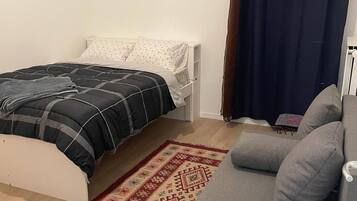 A compact room with a bed, a grey sofa, a patterned rug, and a dark curtain.