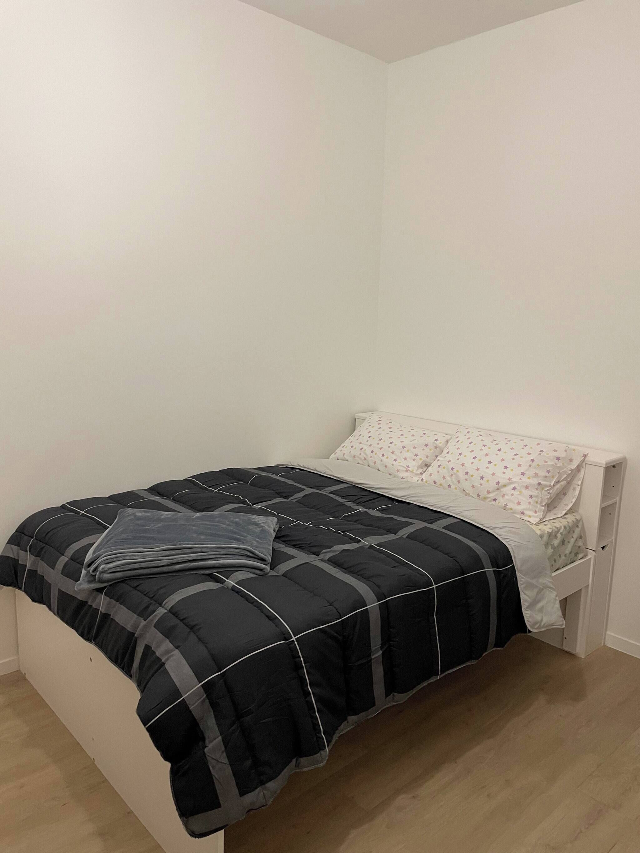 Una cama individual con colcha negra y gris, un almohadón blanco con estampado floral y un cabecero blanco.