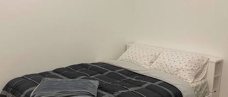 Una cama individual con colcha negra y gris, un almohadón blanco con estampado floral y un cabecero blanco.