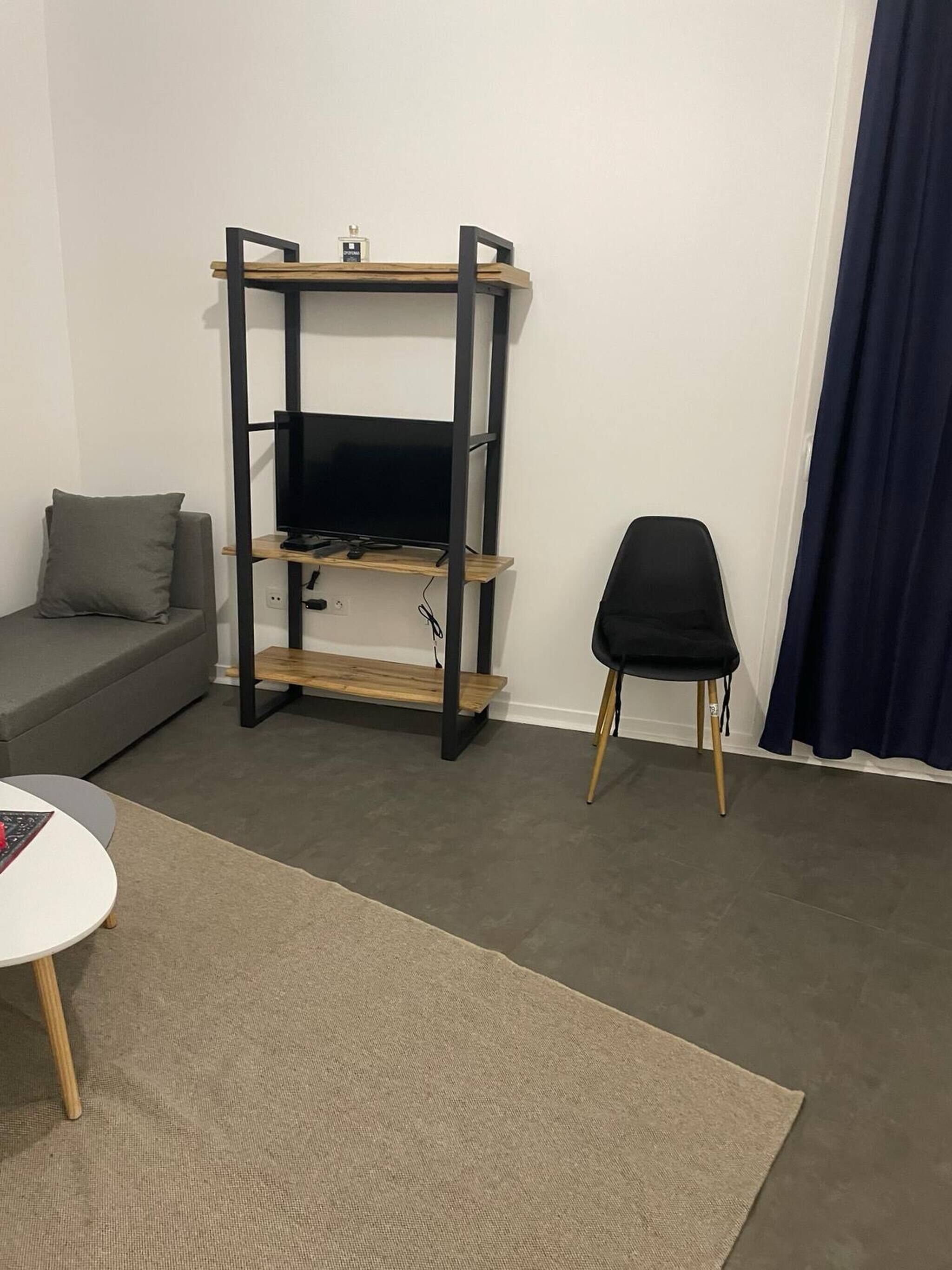 Un salon avec un téléviseur posé sur un support en métal noir, un canapé gris, un fauteuil noir et une petite table ronde.