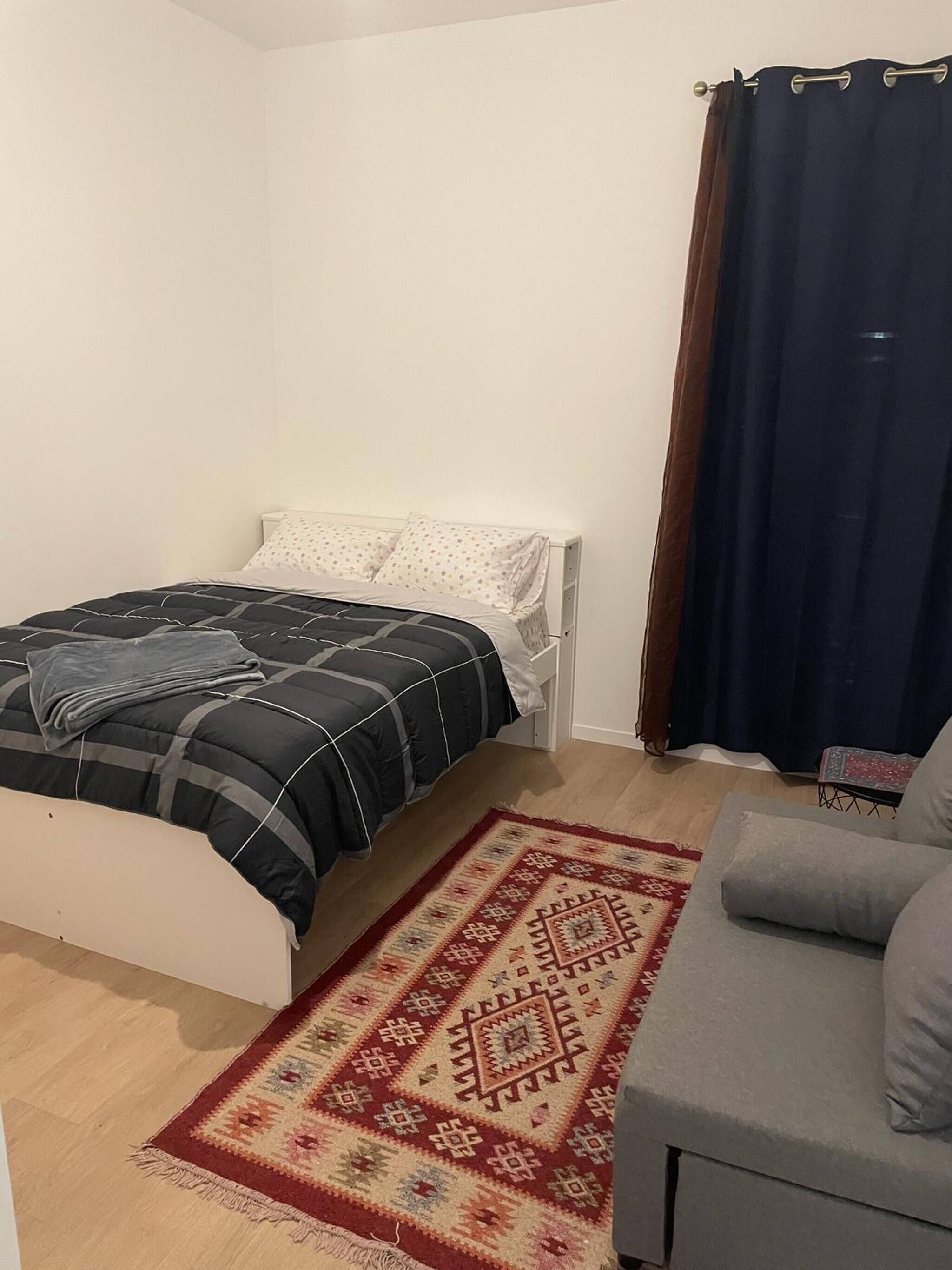 Um quarto com uma cama, um sofá cinzento, um tapete estampado e uma cortina escura.