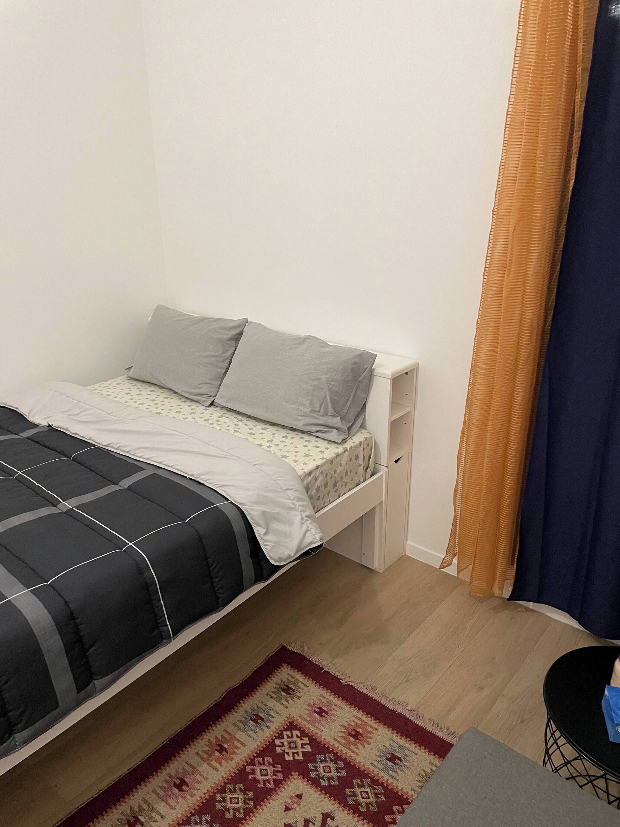 A single bed with a grey and black bedspread, a white bedside table, a small drawer, a patterned rug, and a blue curtain.