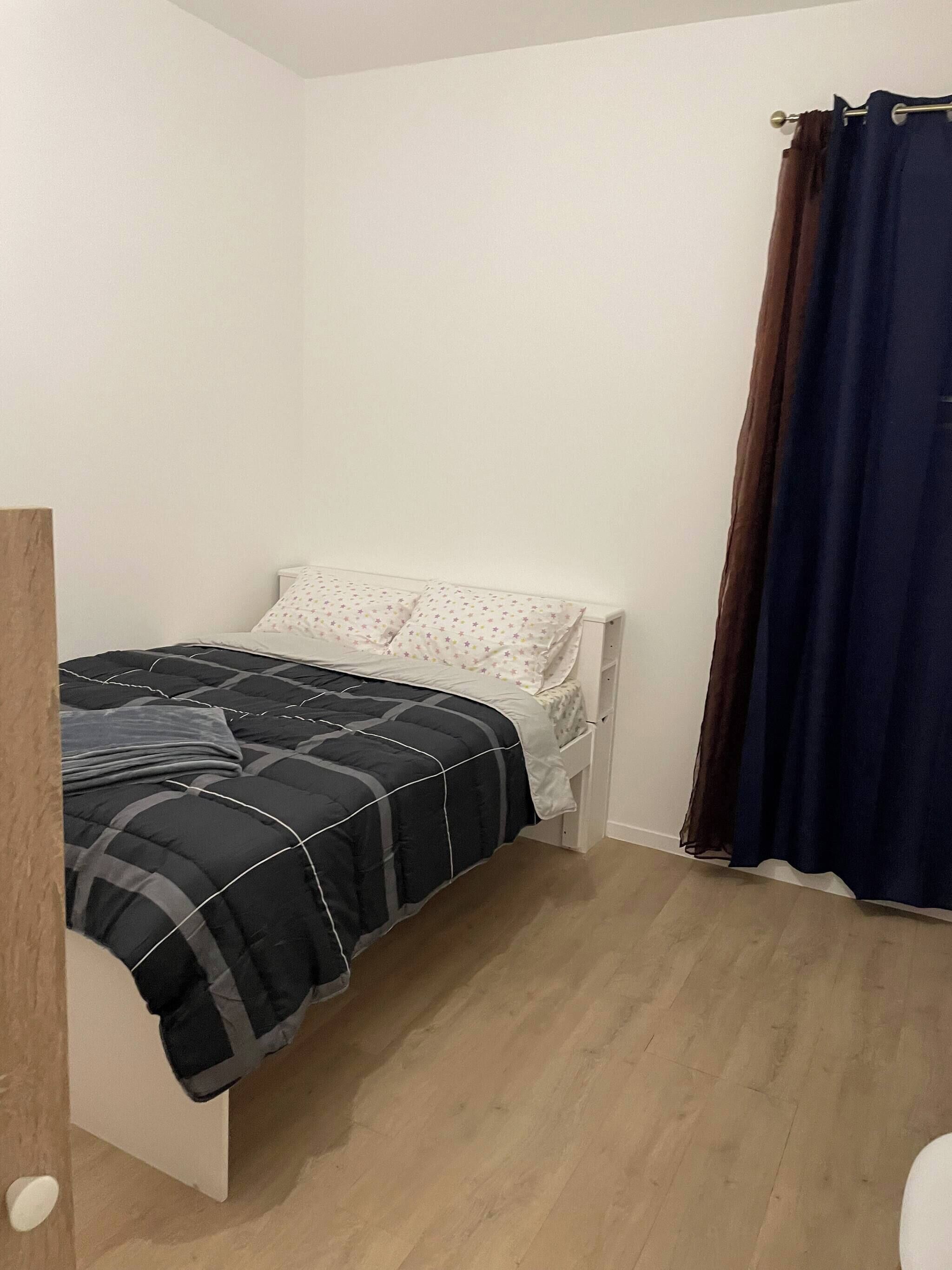 Una habitación pequeña con una cama individual, piso de madera, paredes blancas y una cortina azul oscuro.