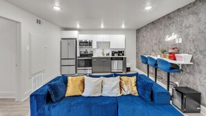 A modern living room with a blue sofa, a kitchen in the background, and a neon sign on the wall. - Renovated 2BR - Sleeps 6 - Walk to Deep Ellum/DT (Dallas)