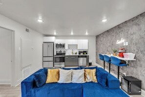 A modern living room with a blue sofa, a kitchen in the background, and a neon sign on the wall. - Renovated 2BR - Sleeps 6 - Walk to Deep Ellum/DT (Dallas)
