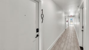 Käytävän seinissä on valkoiset ovet, ja jokaisessa ovessa on numero. Käytävän lattia on laatoitettu ja katossa on upotetut valaisimet.