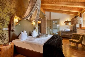 A bedroom with a large bed, a canopy, a wooden headboard, a bedside table, a chair, and a bathroom visible in the background. - Chalet Mi Amur (Badia)