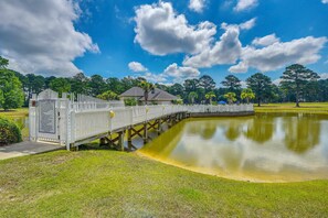 A wooden bridge over a calm pond with a white fence and a signboard, surrounded by greenery and trees. - Gorgeous Ground Floor Condo Just Steps to Pool! (Calabash)