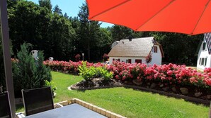 A patio area with a table and chairs, a red umbrella, and a garden with pink flowers and a white house in the background. - Near the beach & newly renovated: Vacation rental at Küstenwaldhaus Koserow - (Koserow)