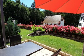A patio area with a table and chairs, a red umbrella, and a garden with pink flowers and a white house in the background. - Near the beach & newly renovated: Vacation rental at Küstenwaldhaus Koserow - (Koserow)