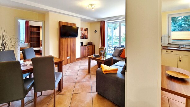 A modern living room with a sofa, dining table, chairs, a TV, and a kitchen area with a window. - Near the beach & newly renovated: Vacation rental at Küstenwaldhaus Koserow - (Koserow)