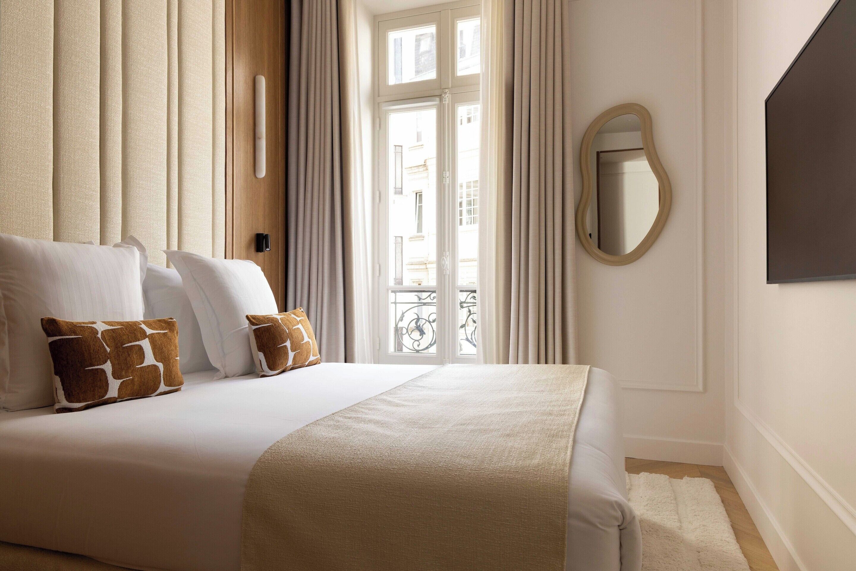 A hotel room with a bed, curtains, a mirror, and a view of the outside.