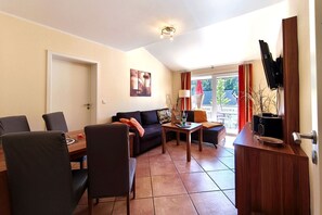 Ein Wohnzimmer mit einer Couch, Sesseln, einem Couchtisch, einem Fernseher und einem Balkon mit roten Vorhängen.