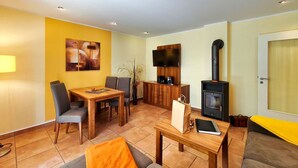 A furnished living room with a dining area, a flat-screen TV, a wood-burning stove, and a door with a window. - Möwe Vacation Rental – Relaxation Between the Forest and the Baltic Sea - Küstenwaldhaus (Koserow)