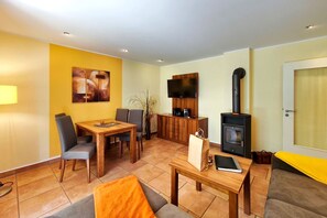 A furnished living room with a dining area, a flat-screen TV, a wood-burning stove, and a door with a window. - Möwe Vacation Rental – Relaxation Between the Forest and the Baltic Sea - Küstenwaldhaus (Koserow)