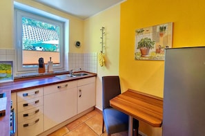 A compact kitchen with a sink, cabinets, a window with blinds, a clock, and a small table with a chair. - Möwe Vacation Rental – Relaxation Between the Forest and the Baltic Sea - Küstenwaldhaus (Koserow)