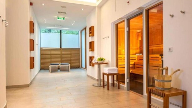 A hotel corridor with a reception desk, a small table with a bowl of fruit, a wooden bench, and a sauna visible through the glass doors.