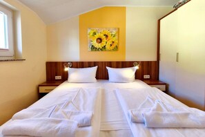 A double bed with white linens, two bedside tables, a wall-mounted painting of sunflowers, and a window with blinds. - Küstenwaldhaus "Muschel" Vacation Rental – Relaxation Between the Forest and the Baltic Sea - (Koserow)