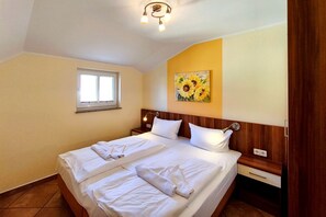 A hotel room with a double bed, a bedside table, a nightstand, a wall-mounted lamp, a window, and a painting of sunflowers. - Küstenwaldhaus "Muschel" Vacation Rental – Relaxation Between the Forest and the Baltic Sea - (Koserow)