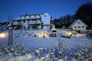 Ein großes, zweistöckiges Haus mit einem mit Schnee bedeckten Rasen und einem beleuchteten Außenofen.
