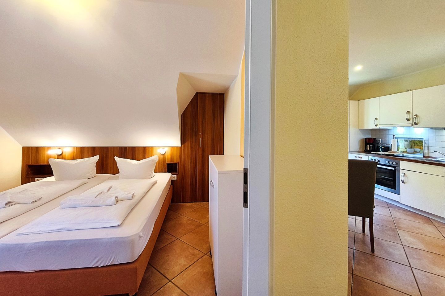 Ein Hotelzimmer mit einem Bett, Nachttischen, einem Holzkopfende, einer kleinen Küchenzeile und einem Sessel.