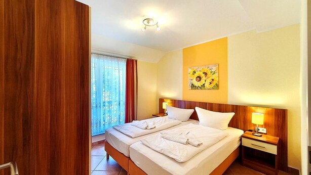 Una cama doble con dos almohadas, una mesita de noche con lámpara, un cuadro de girasoles y vistas a un balcón con cortinas.