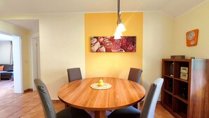 Un comedor con una mesa redonda de madera, cuatro sillas, una pared amarilla con un cuadro, un reloj y una estantería de madera.