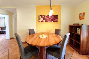 A dining area with a round wooden table, four chairs, a yellow wall with a painting, a clock, and a wooden bookshelf. - Delfin Vacation Rental – Sauna, Fireplace & Near the Beach - Coastal Forest House (Koserow)