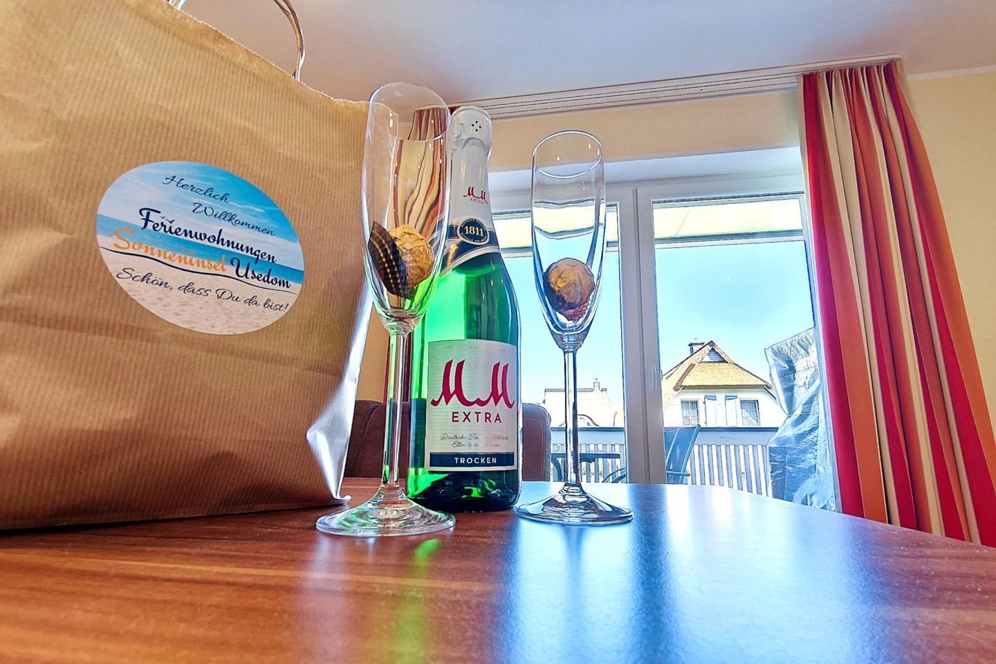 A bottle of 'Müll EXTRA' wine, a glass with a nut, and a paper bag with text on a wooden table by a window.