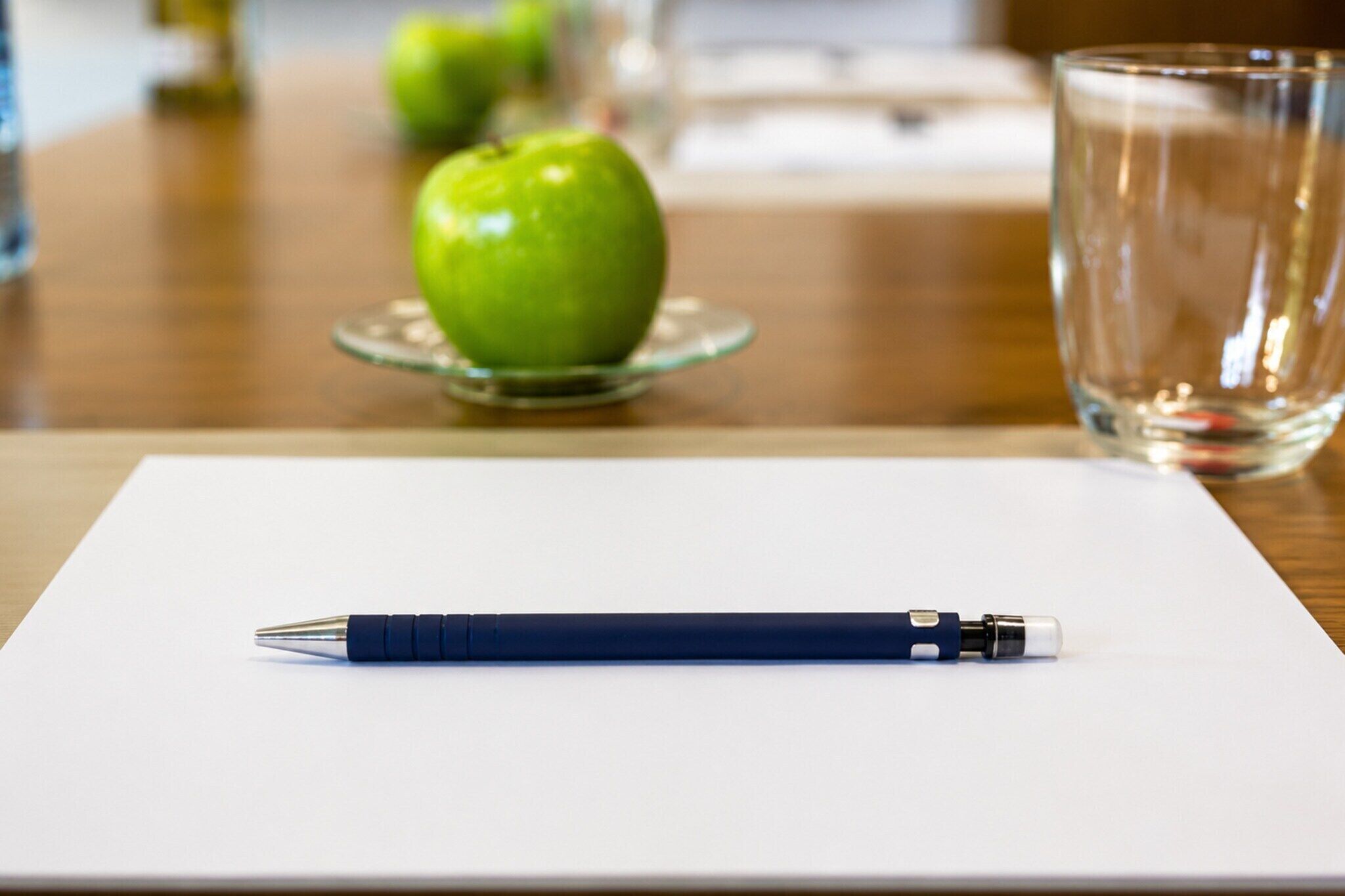 Ein Stift auf einer weißen Oberfläche, im Hintergrund ein Glas und ein Apfel.