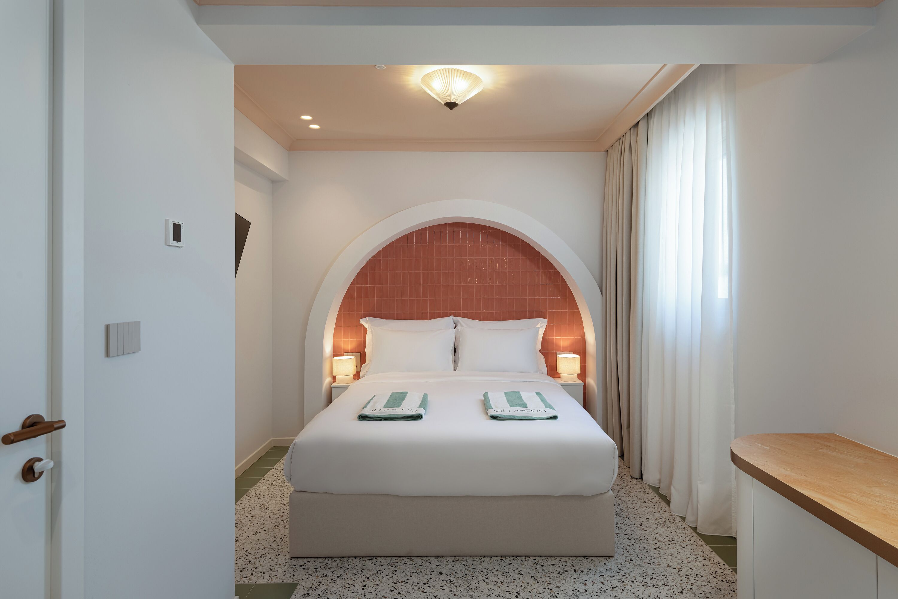 Een hotelkamer met een bed, twee bedlampen, een deur en een klein raam met gordijnen.