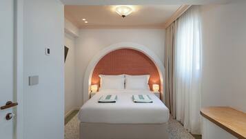 Een hotelkamer met een bed, twee bedlampen, een deur en een klein raam met gordijnen.