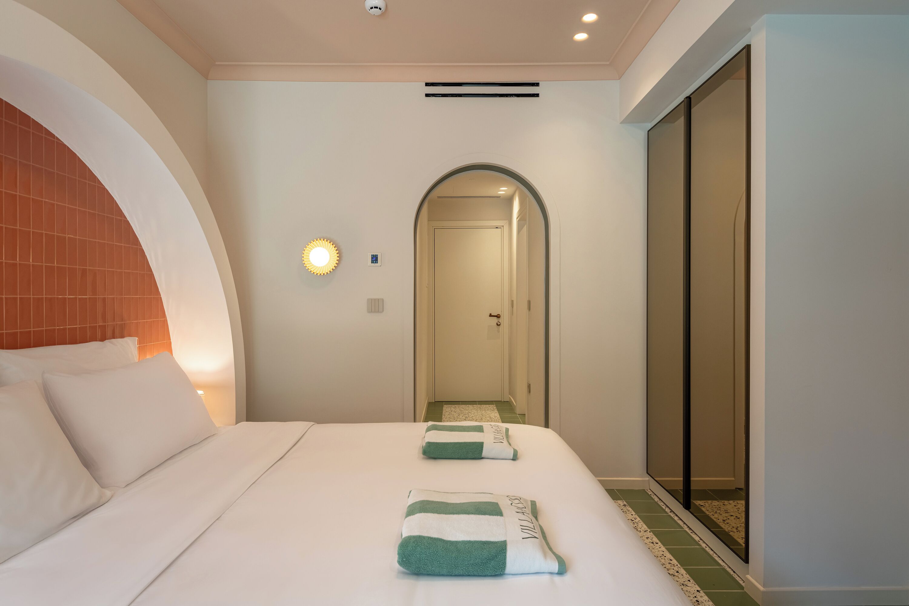 Quarto de hotel com cama, mesa de cabeceira, espelho e porta de acesso a um banheiro.