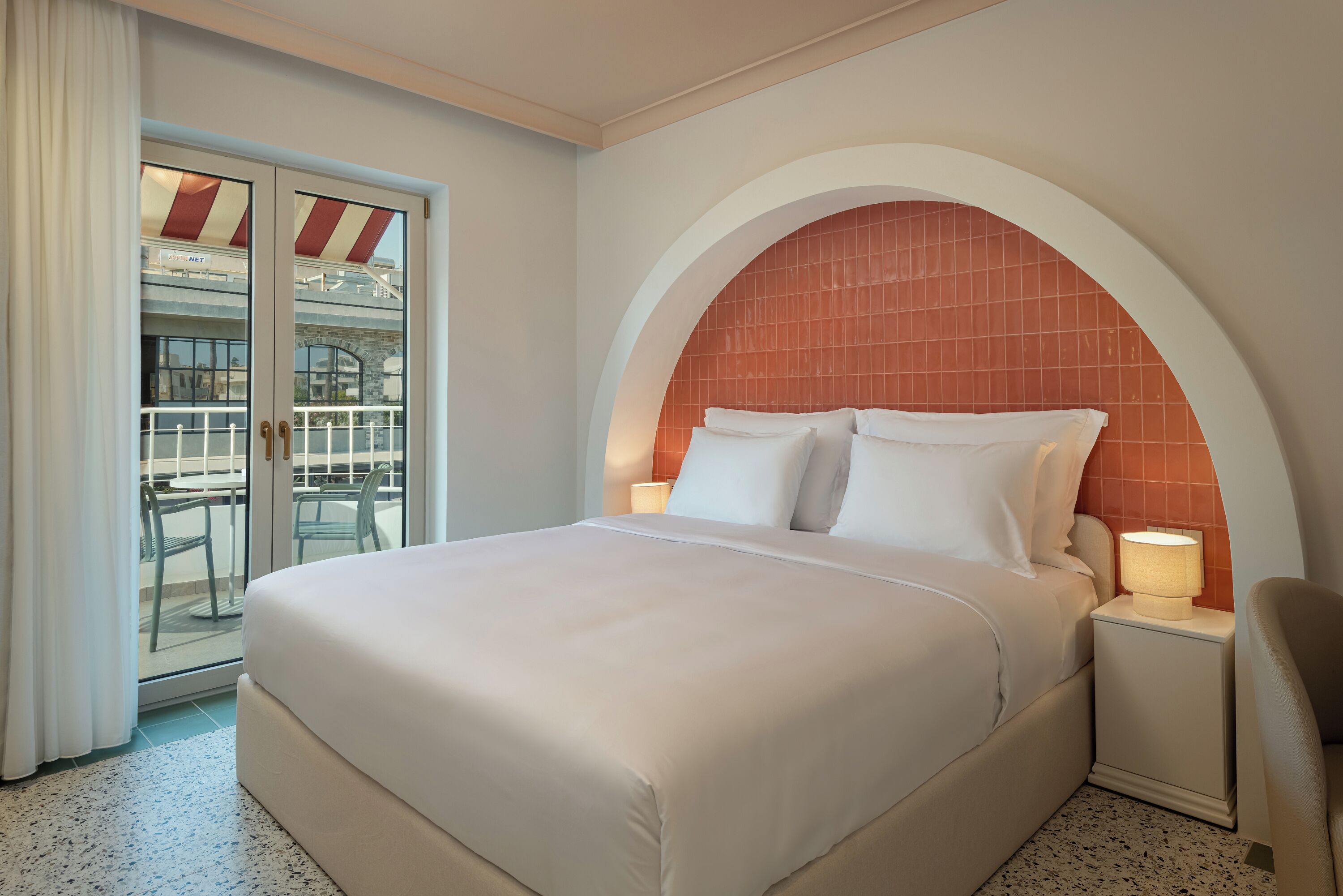 A hotel room with a large bed, a nightstand with a lamp, and a view of a balcony with outdoor furniture.