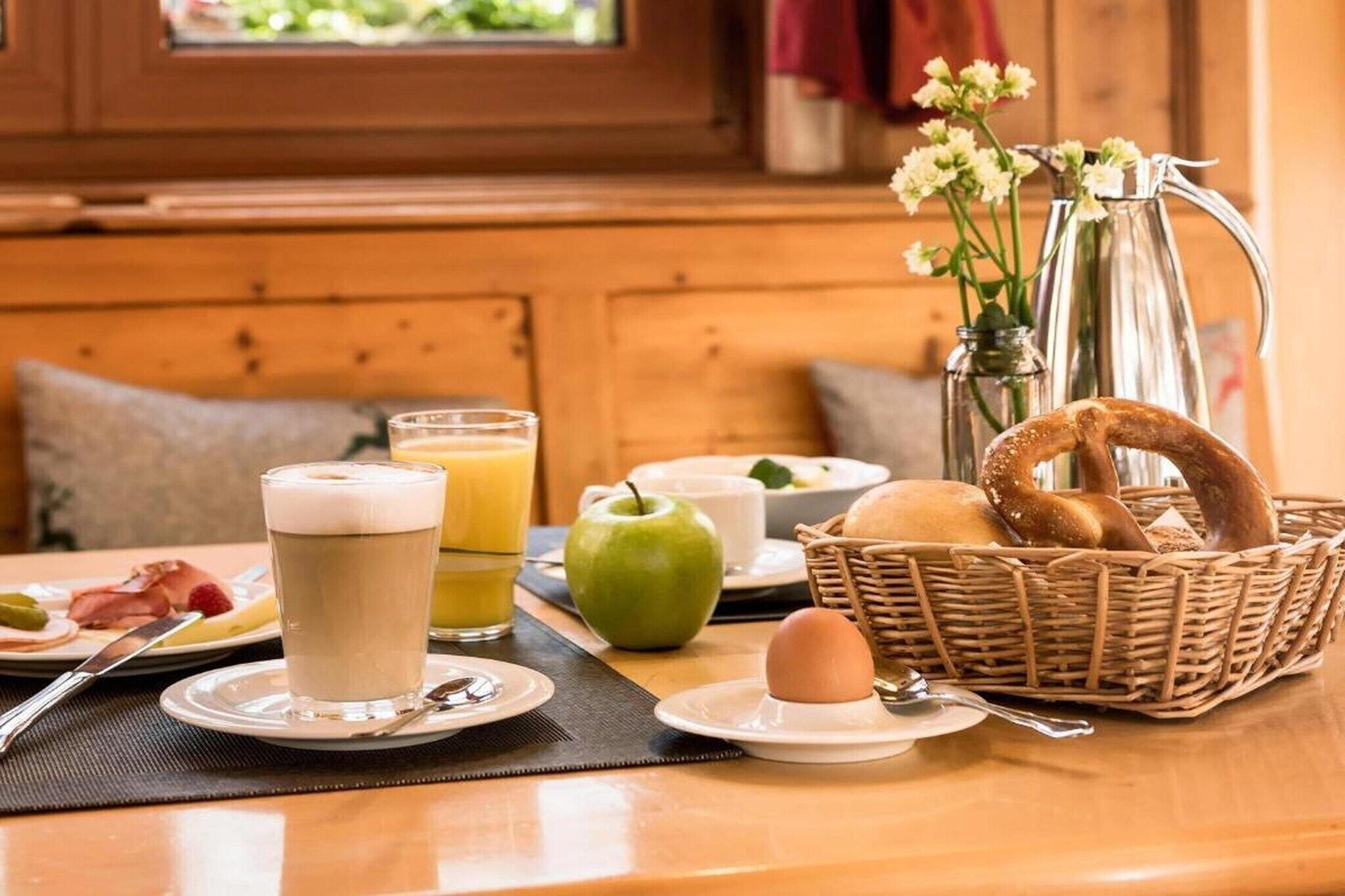 Une table mise pour le petit-déjeuner avec un verre de café, un verre de jus d’orange, une pomme, un panier de pain et une carafe d’eau.