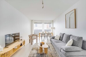 Ein modernes Wohnzimmer mit einem Flachbildfernseher, einem grauen Sofa, einem Esstisch mit Stühlen und einem großen Fenster.
