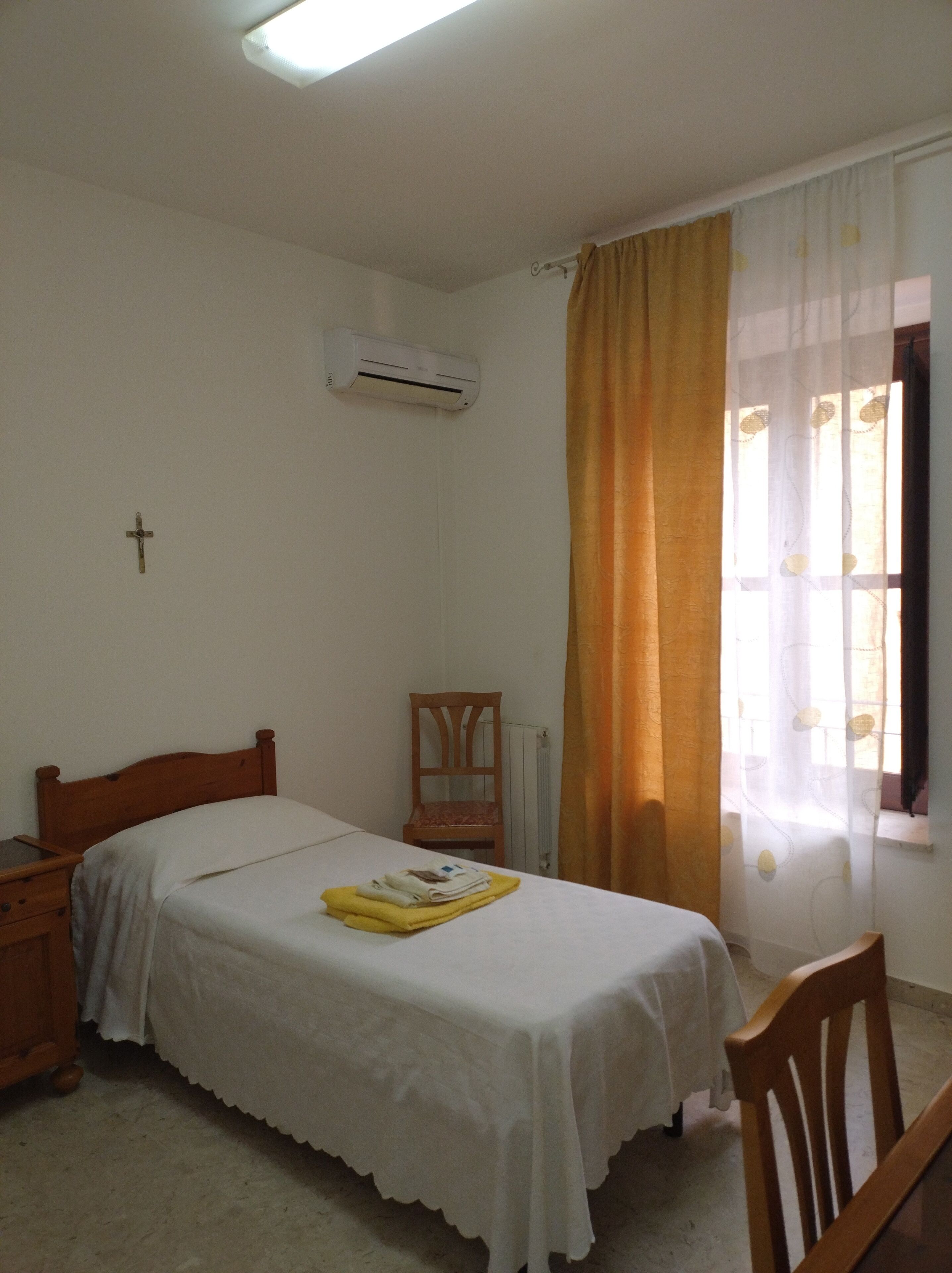 A bedroom with a single bed, a wooden chair, a bedside table, and a window with curtains.