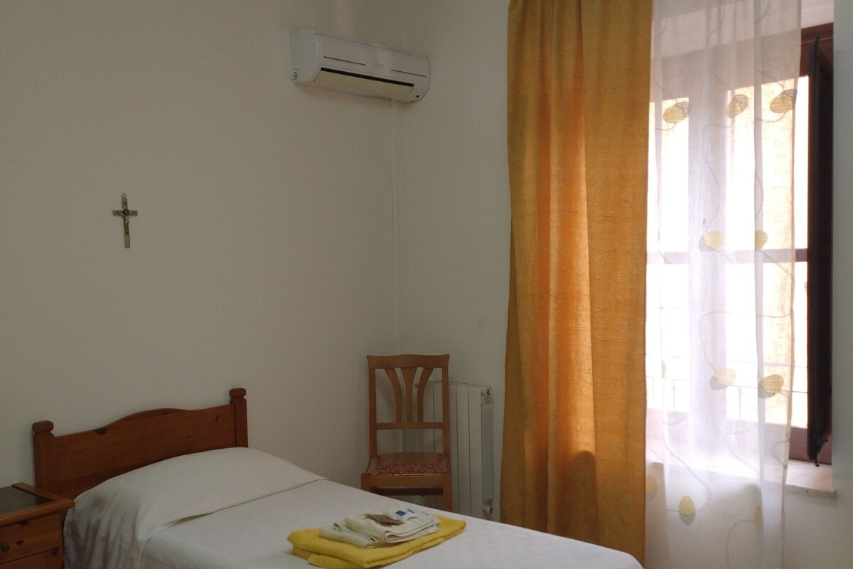 Un dormitorio con una cama individual, una silla de madera, una mesita de noche y una ventana con cortinas.