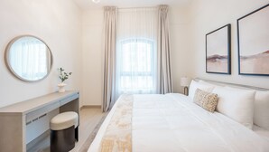Una camera d'hotel con un letto, una scrivania, una sedia, uno specchio rotondo, una finestra con tende e due quadri appesi al muro.