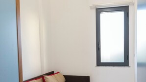 Ein kleines Zimmer mit einem Einzelbett, einem Fenster, einer Tür und Fliesenboden.