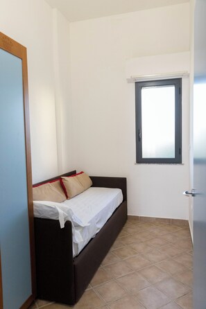 Ein kleines Zimmer mit einem Einzelbett, einem Fenster, einer Tür und Fliesenboden.