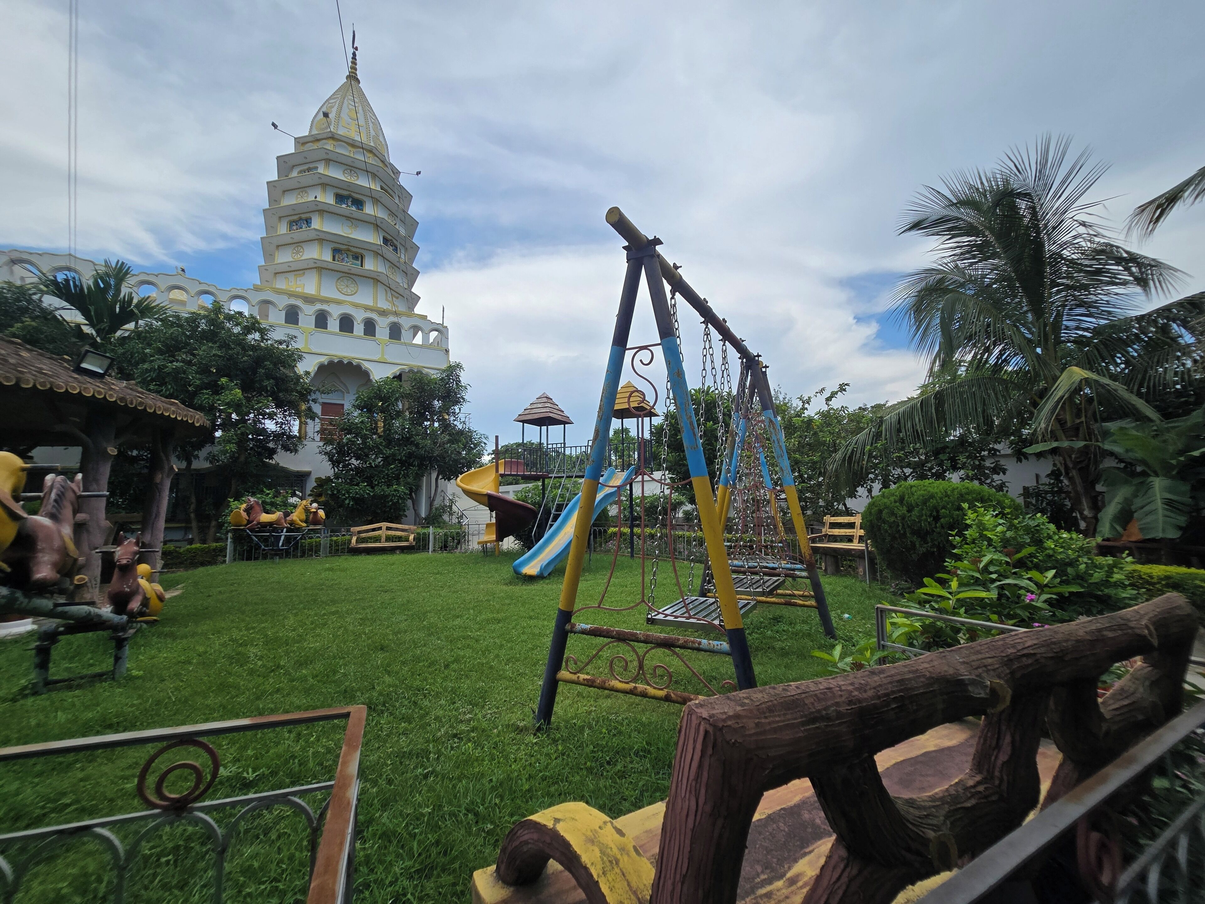 Un parque infantil con columpios y tobogán, rodeado de vegetación y un edificio grande con una torre.