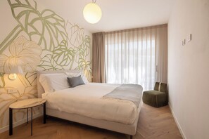 Ein Schlafzimmer mit einem Bett, einem Sessel, einer Lampe, einem Fenster mit Vorhängen und einer Wand mit Pflanzenmuster.