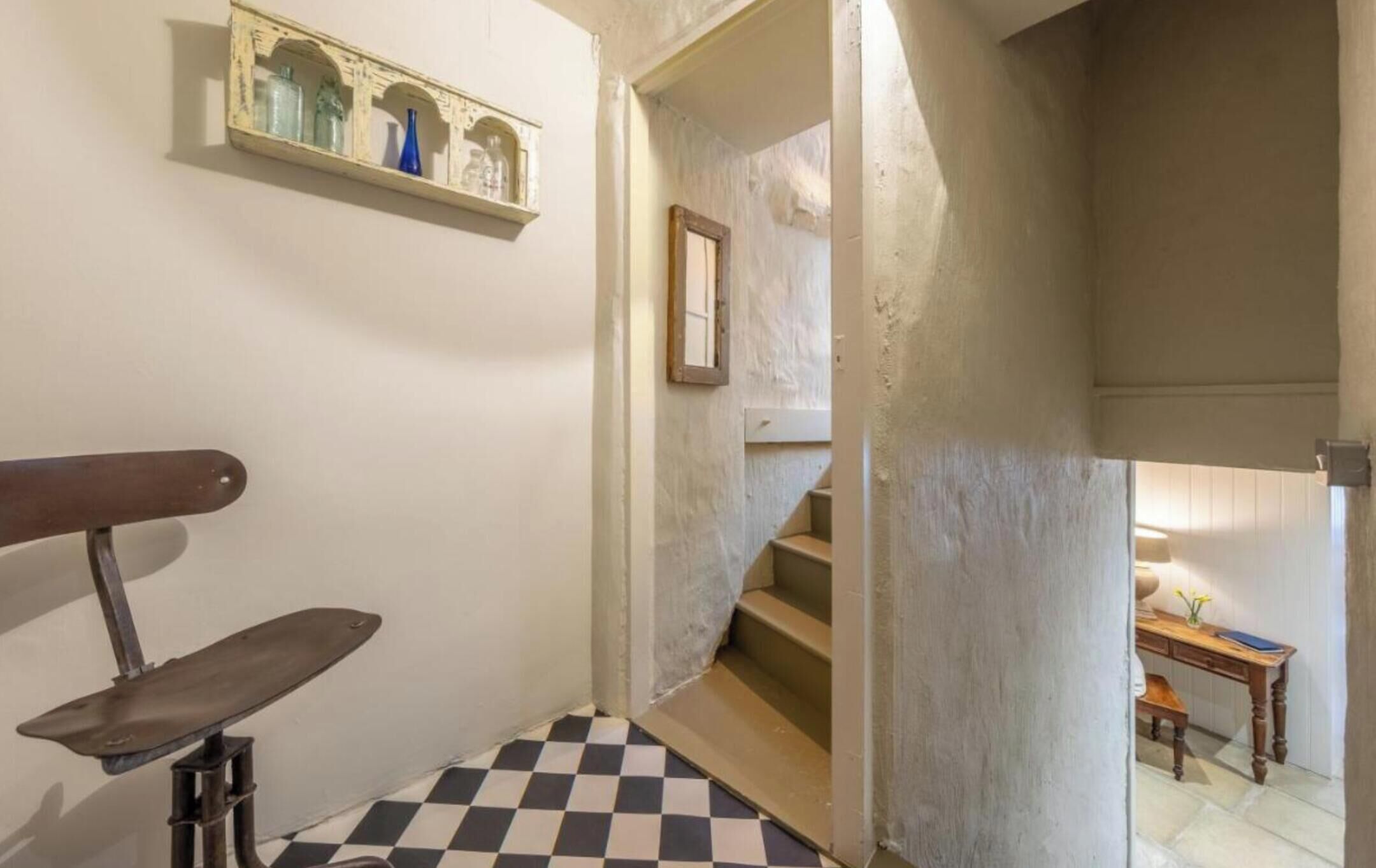 A narrow hallway with a checkered floor, a wooden bench, a shelf with decorative items, and a staircase leading to another room.