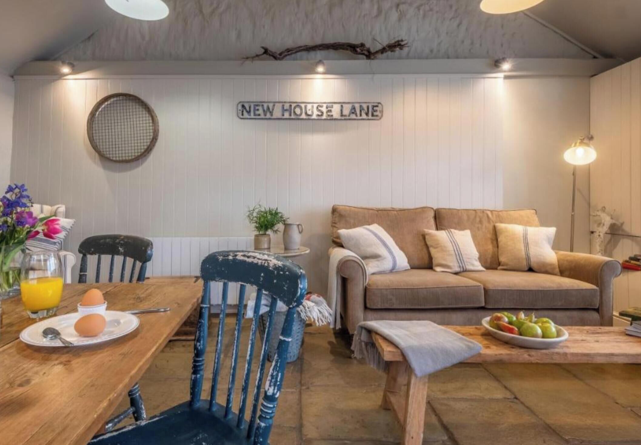 Un salon chaleureux avec une table à manger en bois, une chaise bleue, un canapé marron et une pancarte indiquant « NEW HOUSE LANE ».