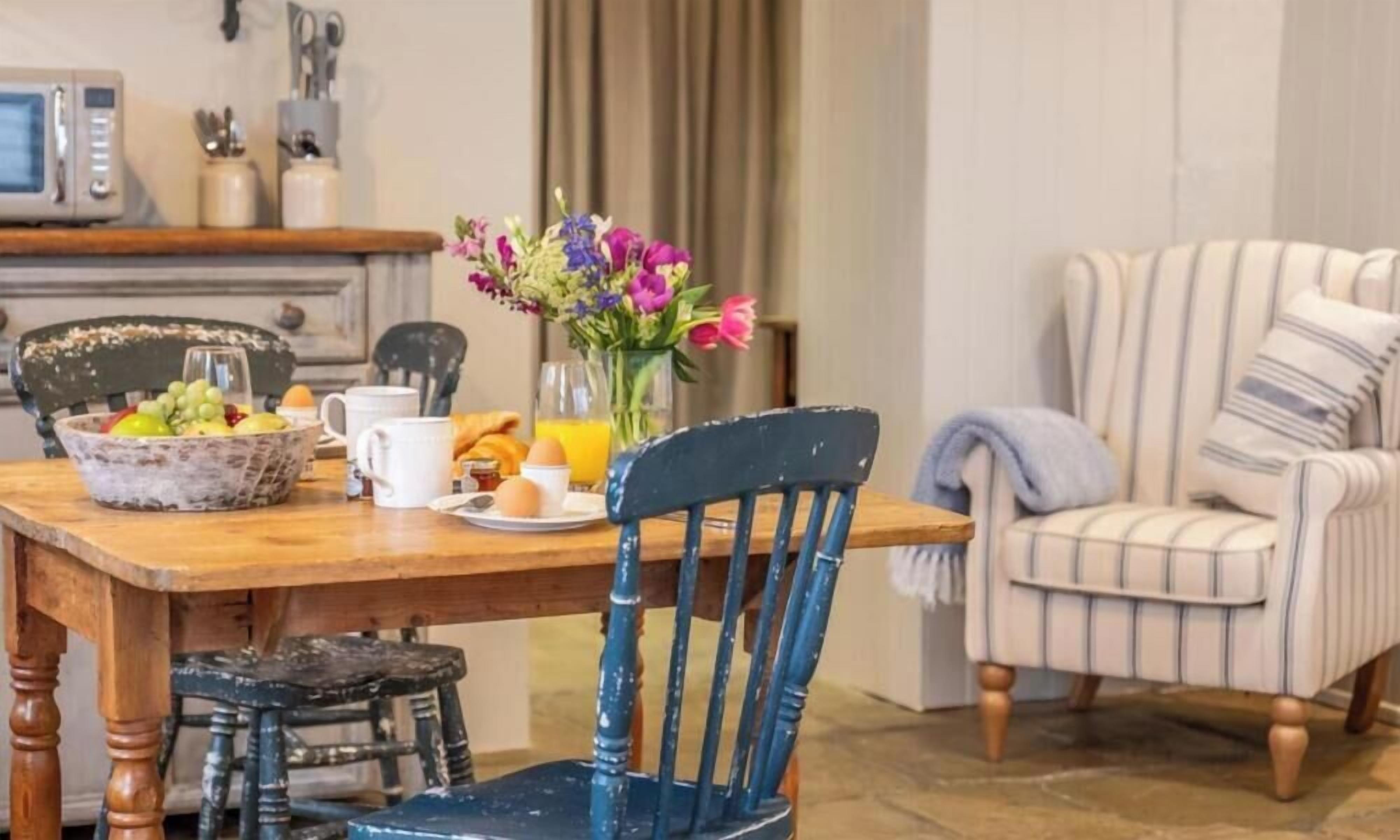 Un comedor con una mesa de madera, sillas, un cuenco de fruta, un florero y un sillón a rayas.