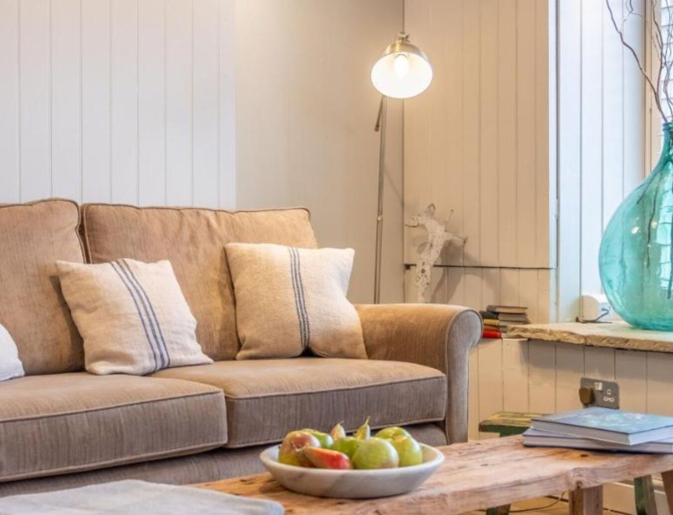 A living room with a sofa, pillows, a bowl of fruit, a lamp, and a vase.